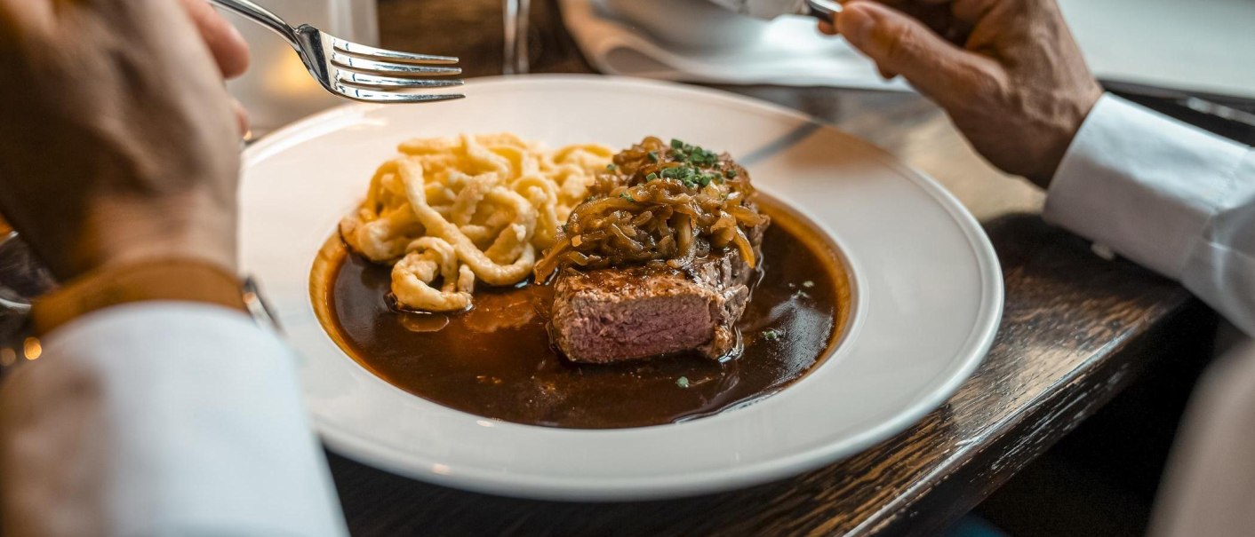Ein Teller mit Steak, Zwiebeln und Spätzle in Soße. Eine Person hält eine Gabel und ein Messer, bereit zum Essen., © SMG, Martina Denker