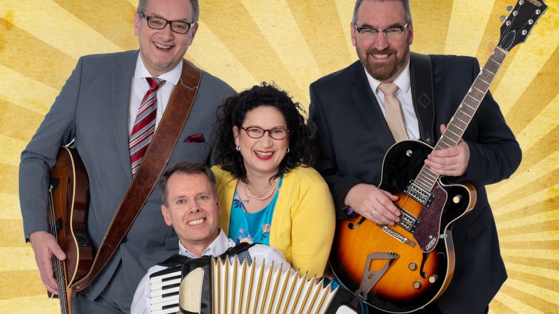 Vier Personen posieren fröhlich mit Instrumenten vor einem gelben Strahlenhintergrund. Zwei Männer halten Gitarren, einer ein Akkordeon, eine Frau steht in der Mitte., © Friedrichsdbau Varieté Vier Personen posieren fröhlich mit Instrumenten vor einem gelben Strahlenhintergrund. Zwei Männer halten Gitarren, einer ein Akkordeon, eine Frau steht in der Mitte., © Friedrichsdbau Varieté
