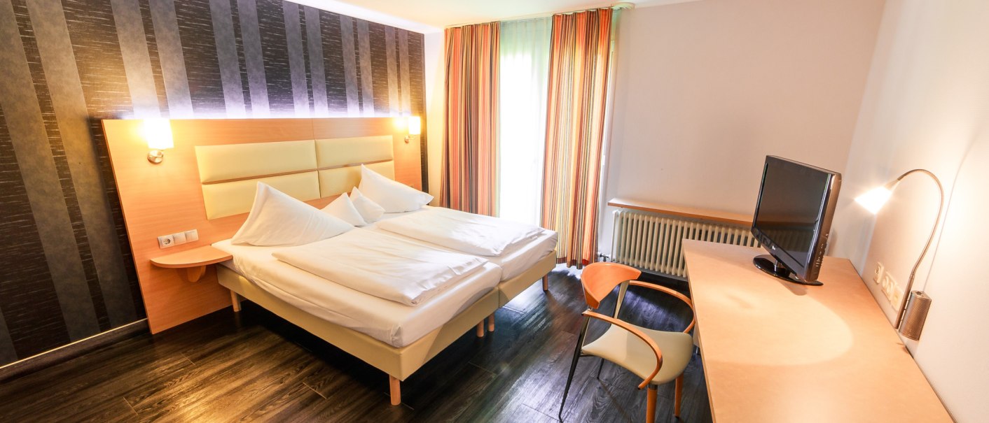 Modern double room with large bed, desk, chair and TV. The walls are partly dark striped, curtains in warm colors., © PLAZA Hotelgroup GmbH