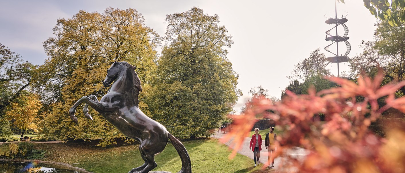 Bronzestatue eines aufsteigenden Pferdes im Höhenpark Killesberg, umgeben von herbstlichen Bäumen und einem spiralförmigen Aussichtsturm im Hintergrund., © SMG, Christoph Düpper Bronzestatue eines aufsteigenden Pferdes im Höhenpark Killesberg, umgeben von herbstlichen Bäumen und einem spiralförmigen Aussichtsturm im Hintergrund., © SMG, Christoph Düpper
