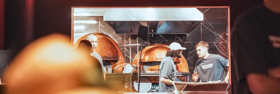 Neon sign reading &ldquo;STRAIGHT OUTTA NAPLES&rdquo; above an open kitchen; three people are working in the kitchen, presumably at a L&rsquo;Artista grand opening., &copy; L'Artista, Stuttgart
