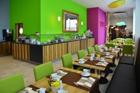 Ein modernes Café mit bunten Wänden, Holztischen und grünen Stühlen. Eine Theke mit Speisen und Getränken ist sichtbar., © TOMAS