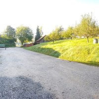 Ländliche Szene mit Holzstapeln an einem Gebäude, einem Anhänger und Obstbäumen auf einer Wiese bei sonnigem Wetter., © Mannschreck-Weine