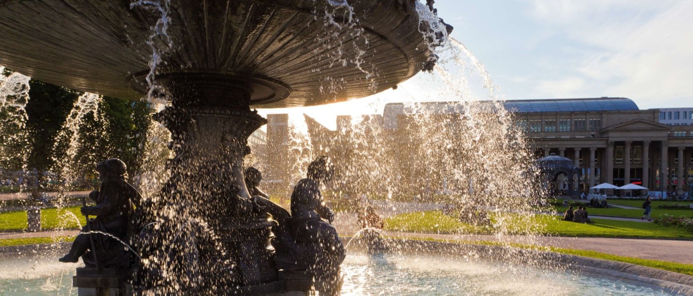 Ein Brunnen am Schlossplatz mit sprudelndem Wasser im Vordergrund. Im Hintergrund sind historische Gebäude und Menschen auf einer Wiese zu sehen., © Werner Dieterich