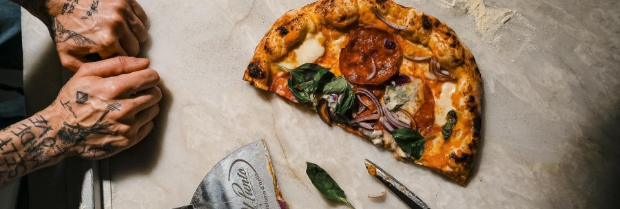 A half-eaten pizza with basil and onions on a light-colored stone, with a pizza cutter and scissors next to it; on the left, two tattooed hands., &copy; L'Artista, Stuttgart
