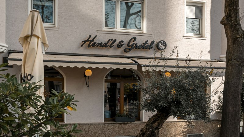Das Bild zeigt die Fassade des Restaurants Hendl & Gretel mit einem Sonnenschirm und Pflanzen im Vordergrund., &copy; SMG, Sarah Schmid