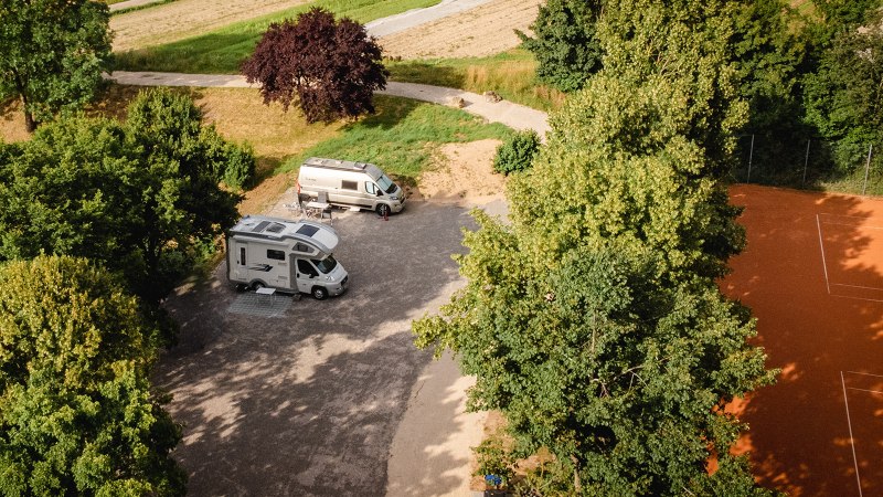 Zwei Wohnmobile stehen auf einem Stellplatz, umgeben von Bäumen. Ein Tennisplatz ist rechts im Bild zu sehen., © Blu Dolci Artwork Zwei Wohnmobile stehen auf einem Stellplatz, umgeben von Bäumen. Ein Tennisplatz ist rechts im Bild zu sehen., © Blu Dolci Artwork