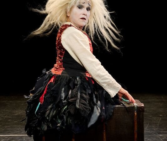 Eine Person mit wildem, blondem Haar und einem auffälligen Kostüm sitzt auf einem Koffer auf einer dunklen Bühne., © Theaterhaus Stuttgart e.V. Eine Person mit wildem, blondem Haar und einem auffälligen Kostüm sitzt auf einem Koffer auf einer dunklen Bühne., © Theaterhaus Stuttgart e.V.