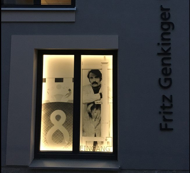 Beleuchtetes Fenster mit einem Bild eines Mannes und der Aufschrift 'Eingang'. An der Außenwand steht 'Fritz Genkinger'., © Freundeskreis Fritz Genkinger e.V.