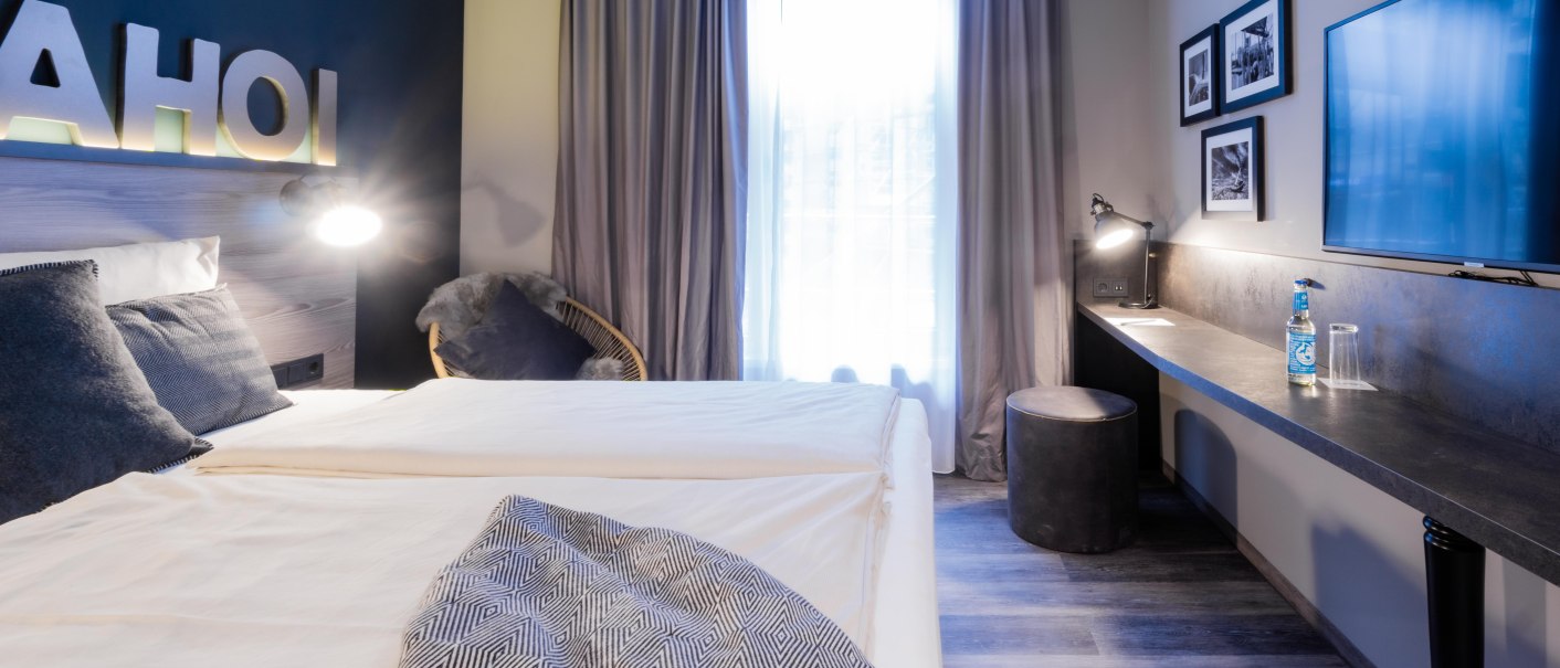 Modernes Zimmer mit Doppelbett, Schreibtisch, Flachbildfernseher und Dekoration. Schriftzug 'AHOI' an der Wand, neutrale Farben und gem&uuml;tliche Atmosph&auml;re., &copy; HARBR. hotel Ludwigsburg / DQuadrat Living GmbH