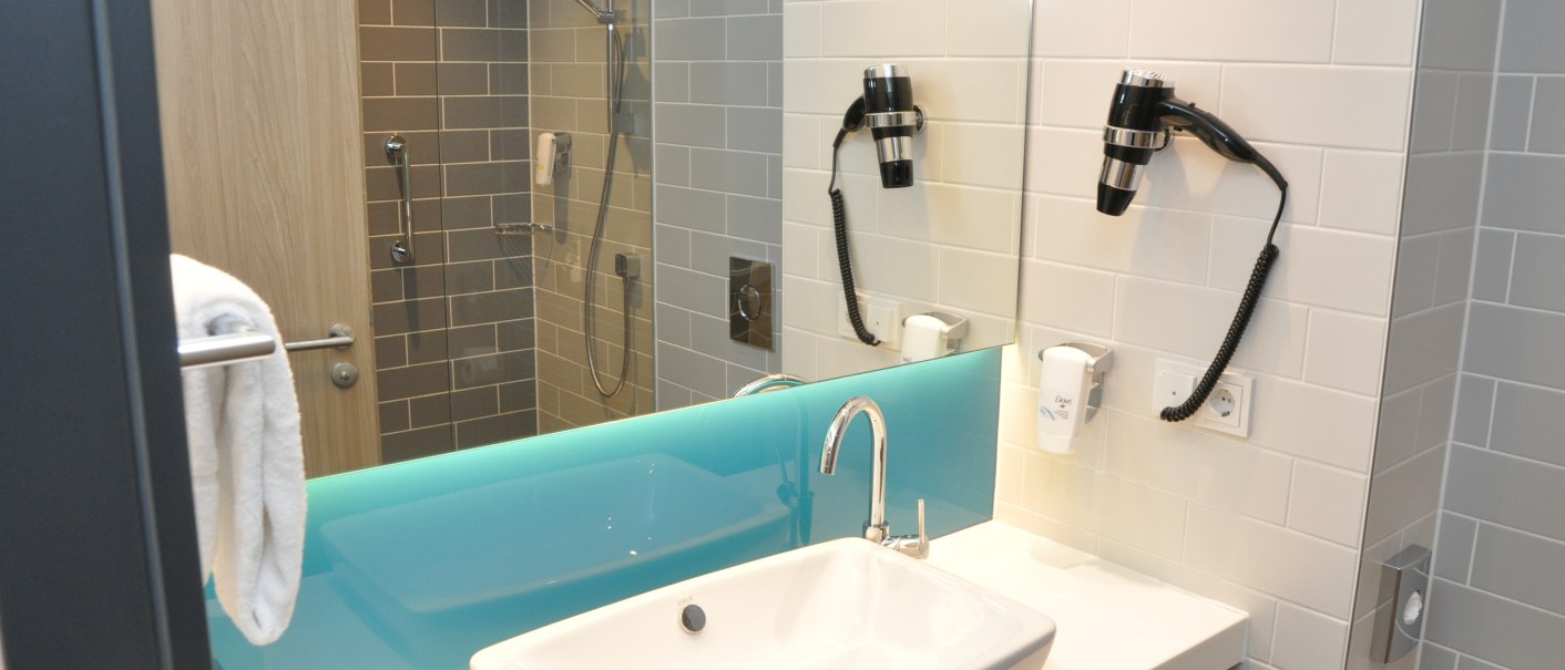 Modernes Badezimmer im Holiday Inn Express Sindelfingen mit Waschbecken, Spiegel, Föhn und Dusche. Helle Fliesen und ein blaues Glasdetail., © tristar GmbH Modernes Badezimmer im Holiday Inn Express Sindelfingen mit Waschbecken, Spiegel, Föhn und Dusche. Helle Fliesen und ein blaues Glasdetail., © tristar GmbH