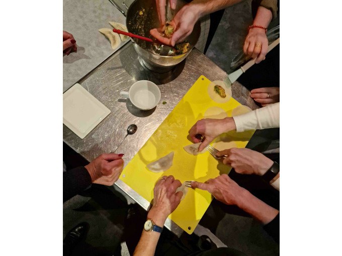 Teilnehmer eines Kochkurses formen gemeinsam Teigtaschen auf einem gelben Schneidebrett. Verschiedene H&auml;nde arbeiten zusammen., &copy; Kultweine-Kochwelten-Kunst&uuml;cke - Kochschule Sonja Lenz