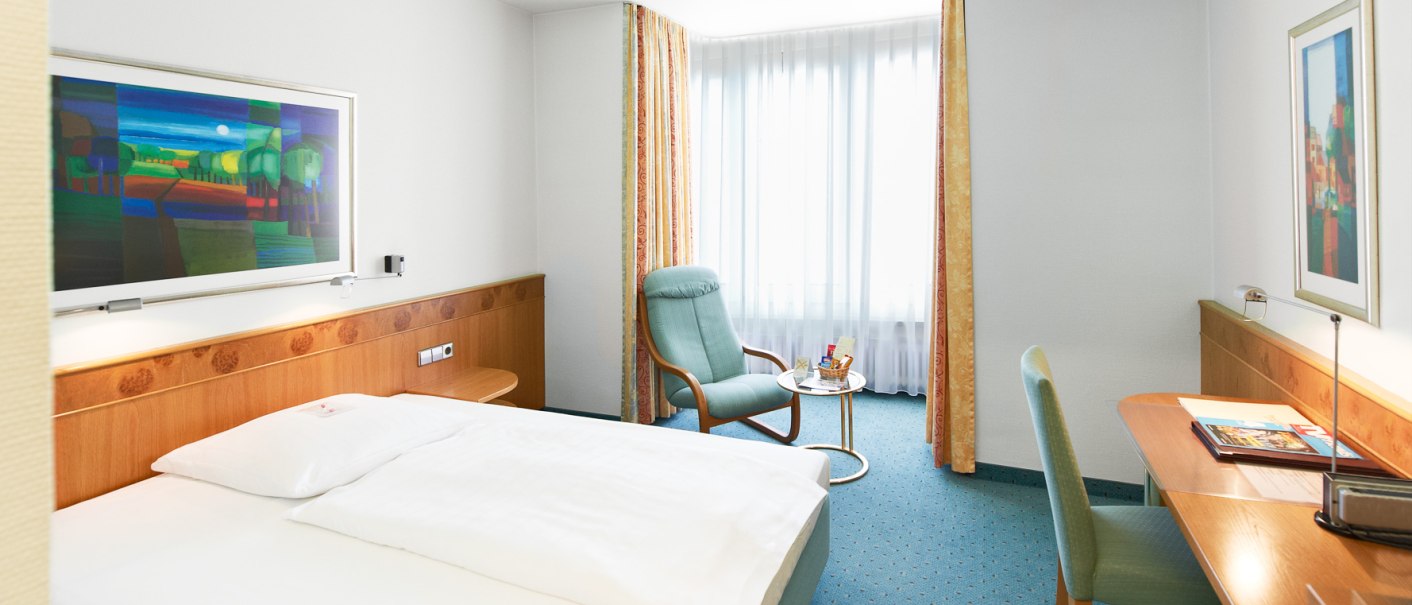 Helles Hotelzimmer mit Doppelbett, Schreibtisch, Sessel und großem Fenster. An der Wand hängt ein buntes Bild., © Hirsch Hotel