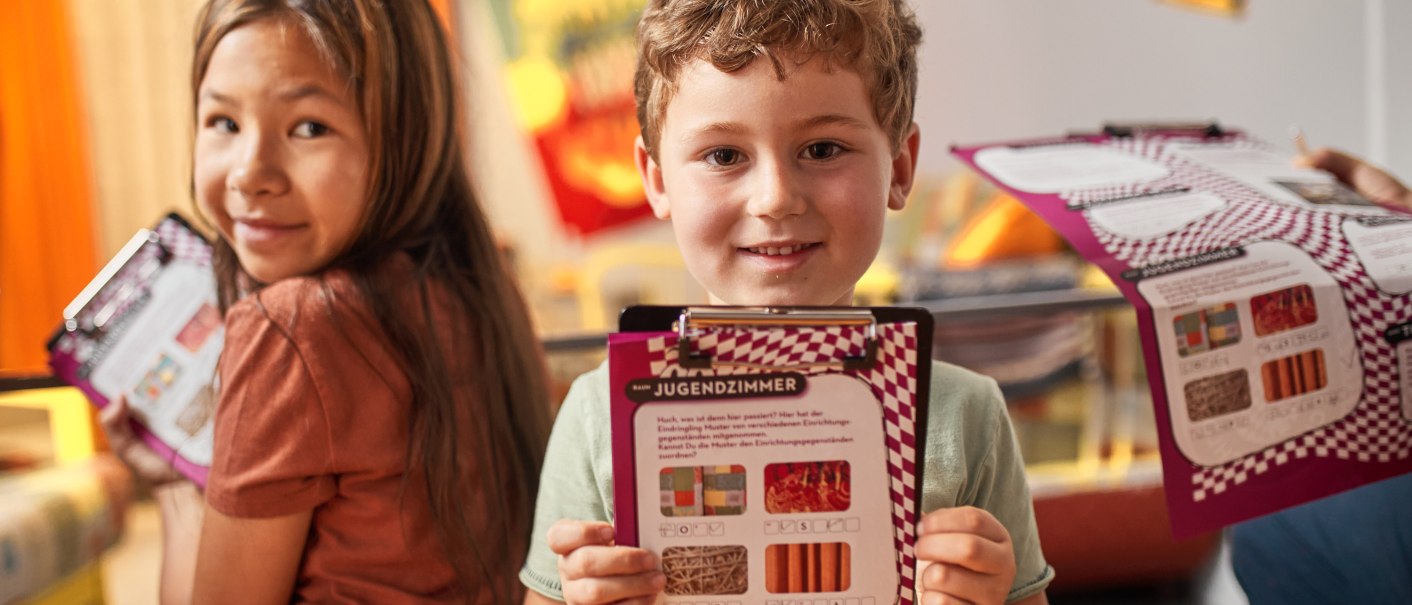 Two children in the Museum of Everyday Culture holding task sheets. They are smiling and appear to be taking part in an activity., © FTGRF.de Two children in the Museum of Everyday Culture holding task sheets. They are smiling and appear to be taking part in an activity., © FTGRF.de