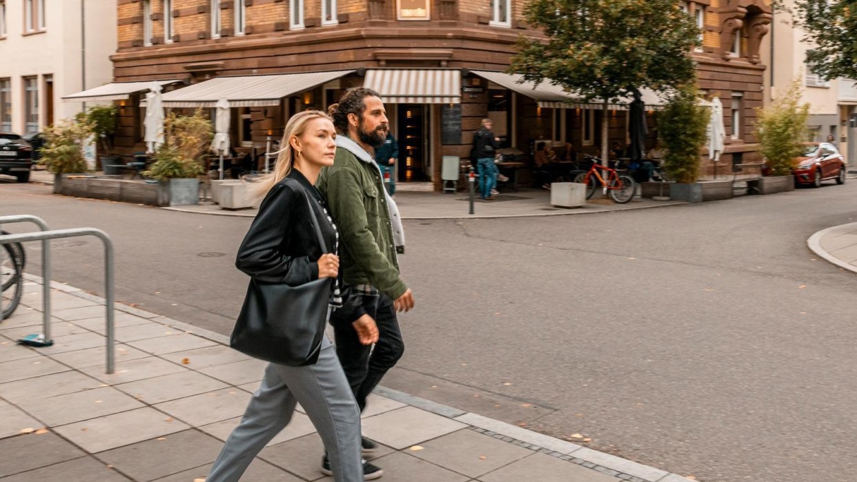 A man and a woman cross a street in an urban environment. A café with outdoor seating can be seen in the background., © SMG, Sarah Schmid A man and a woman cross a street in an urban environment. A café with outdoor seating can be seen in the background., © SMG, Sarah Schmid