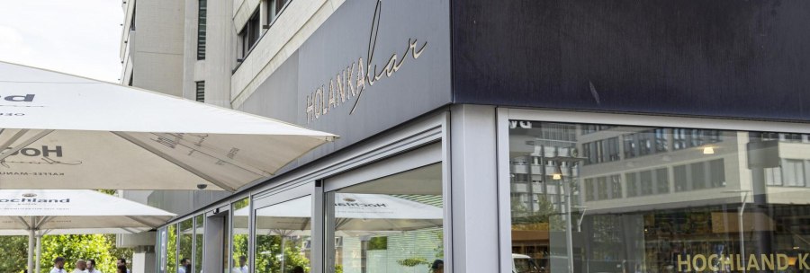 Exterior view of the Holanka Bar with large parasols. People sitting and standing in the outdoor area. Modern buildings in the background., &copy; SMG, Sarah Schmid