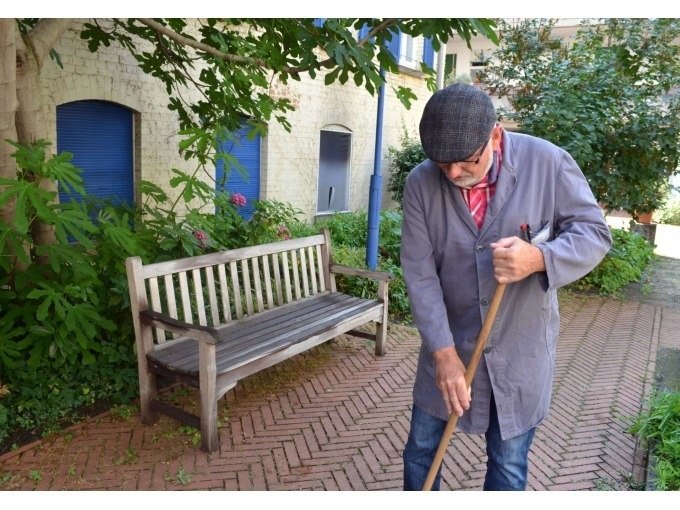 Ein Mann in grauem Kittel fegt einen gepflasterten Weg. Neben ihm steht eine Holzbank, umgeben von Pflanzen und einem Gebäude mit blauen Fensterläden., © Stuttgart Marketing GmbH Ein Mann in grauem Kittel fegt einen gepflasterten Weg. Neben ihm steht eine Holzbank, umgeben von Pflanzen und einem Gebäude mit blauen Fensterläden., © Stuttgart Marketing GmbH