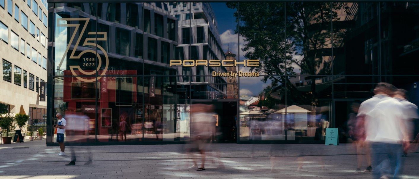 Glasfassade des Porsche Brand Stores mit Jubil&auml;umslogo '75 Jahre 1948-2023'. Menschen gehen vorbei, Geb&auml;ude und B&auml;ume spiegeln sich im Glas., &copy; Porsche AG
