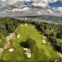 Aerial view of a golf course in Stuttgart, surrounded by trees and hills, with the city in the background under a cloudy sky., © GolfKultur Stuttgart Aerial view of a golf course in Stuttgart, surrounded by trees and hills, with the city in the background under a cloudy sky., © GolfKultur Stuttgart