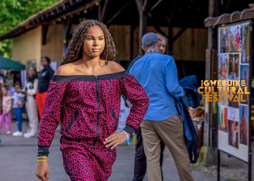 Eine Frau präsentiert ein rotes, gemustertes Outfit auf einem Laufsteg beim Igwe Buike Cultural Festival. Zuschauer im Hintergrund., © igbo Cultural Foundation e.V.