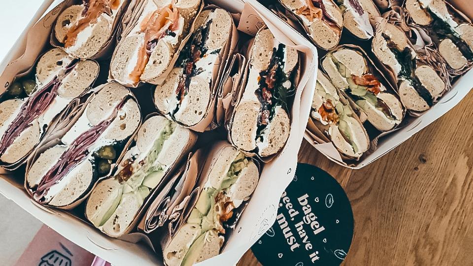 Eine Box mit verschiedenen gefüllten Bagels, darunter Lachs, Salami und Gemüse, auf einem Holztisch. Ein runder Untersetzer ist daneben sichtbar., © Cupcakes & Bagels, Stuttgart