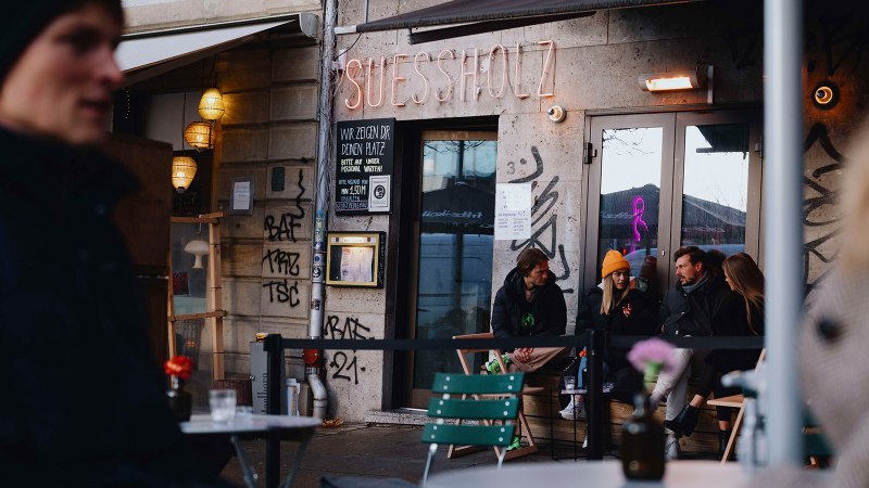 Außenansicht des Cafés 'Suessholz' im Leonhardsviertel. Menschen sitzen draußen, unterhalten sich. Graffiti an den Wänden, Neonlicht im Fenster., © Stuttgart-Marketing GmbH, Alwin Maigler Außenansicht des Cafés 'Suessholz' im Leonhardsviertel. Menschen sitzen draußen, unterhalten sich. Graffiti an den Wänden, Neonlicht im Fenster., © Stuttgart-Marketing GmbH, Alwin Maigler