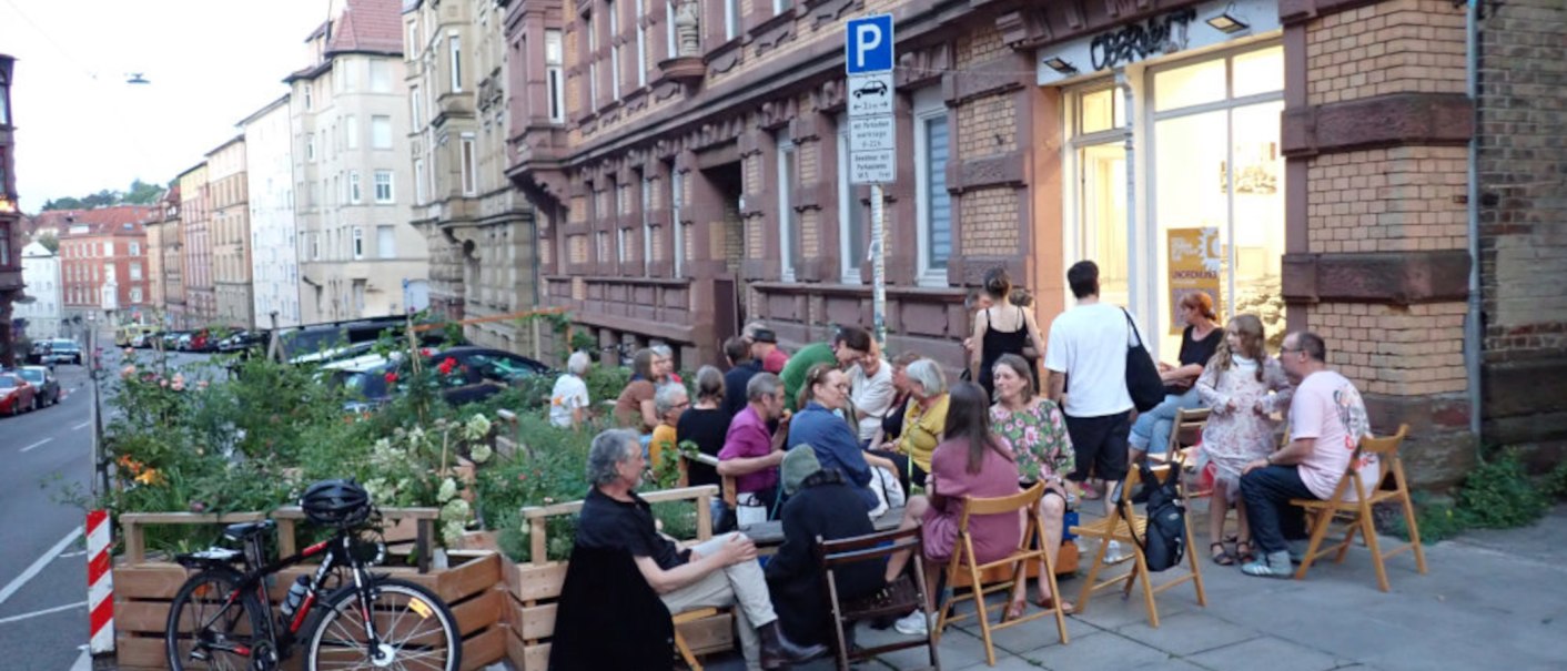 A group of people is sitting on chairs on a sidewalk in an urban setting. Buildings and a bicycle can be seen in the background., © Oberwelt e.V. A group of people is sitting on chairs on a sidewalk in an urban setting. Buildings and a bicycle can be seen in the background., © Oberwelt e.V.