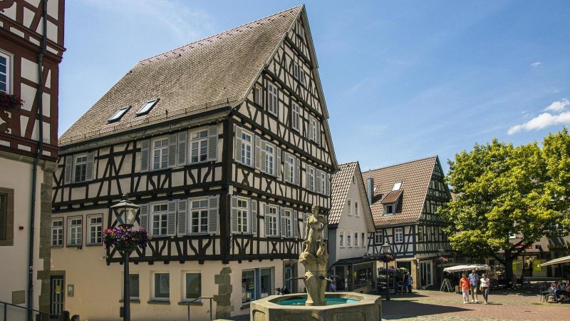 Half-timbered houses on Backnang's market square, with a fountain in the foreground and trees in the background. People stroll and sit in cafés., © Stuttgart-Marketing GmbH, Sarah Schmid Half-timbered houses on Backnang's market square, with a fountain in the foreground and trees in the background. People stroll and sit in cafés., © Stuttgart-Marketing GmbH, Sarah Schmid