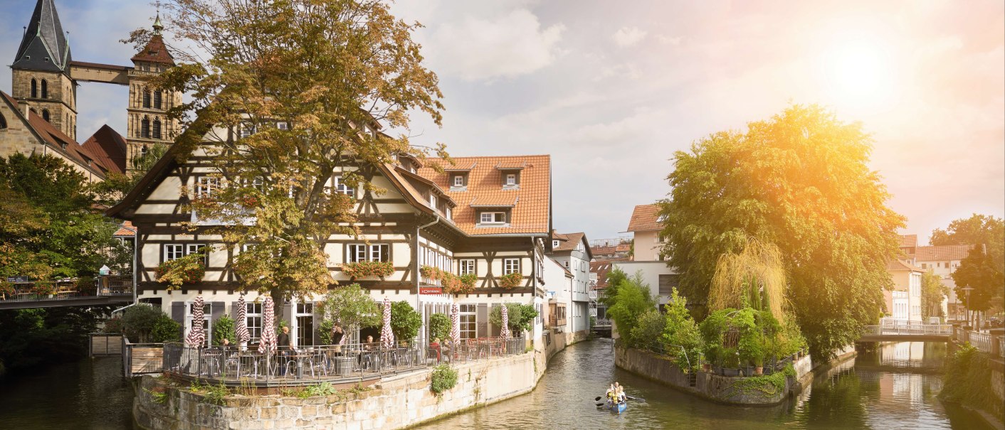 A half-timbered house on the Roßneckar Canal, surrounded by trees. People are sitting in the café while canoeists paddle along the water. Sunshine in the background., © Stuttgart-Marketing GmbH A half-timbered house on the Roßneckar Canal, surrounded by trees. People are sitting in the café while canoeists paddle along the water. Sunshine in the background., © Stuttgart-Marketing GmbH