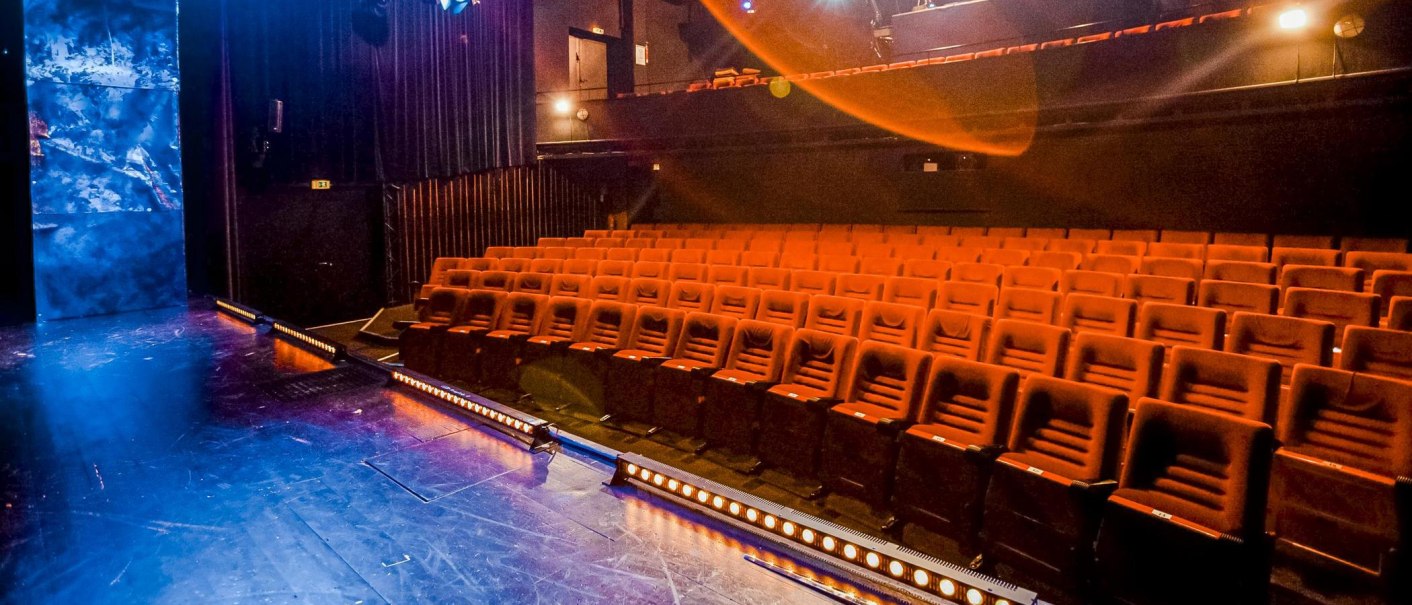 Leerer Theatersaal mit roten Sitzen und einer Bühne, die in blauem Licht erstrahlt. Die Atmosphäre ist ruhig und einladend., © Theater der Altstadt, Stuttgart