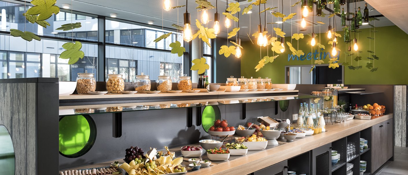 Ein reichhaltiges Frühstücksbuffet mit Obst, Käse, Wurst und Getränken in einem modernen, hellen Raum mit dekorativen Lichtern., © ibis Styles Stuttgart Vaihingen Ein reichhaltiges Frühstücksbuffet mit Obst, Käse, Wurst und Getränken in einem modernen, hellen Raum mit dekorativen Lichtern., © ibis Styles Stuttgart Vaihingen