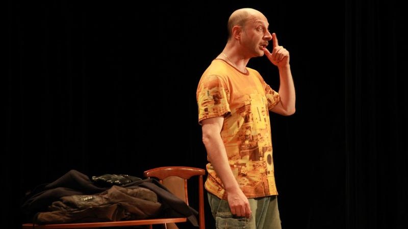 Ein Mann in einem orangefarbenen T-Shirt steht auf einer Bühne und gestikuliert. Neben ihm steht ein Tisch mit Kleidung., © Theater Atelier Ein Mann in einem orangefarbenen T-Shirt steht auf einer Bühne und gestikuliert. Neben ihm steht ein Tisch mit Kleidung., © Theater Atelier