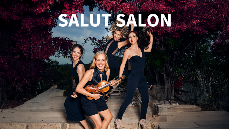 Vier Frauen in schwarzen Outfits posieren fröhlich mit einer Geige vor einer blühenden Kulisse. Oben steht der Schriftzug 'Salut Salon'., © links im Bild Vier Frauen in schwarzen Outfits posieren fröhlich mit einer Geige vor einer blühenden Kulisse. Oben steht der Schriftzug 'Salut Salon'., © links im Bild
