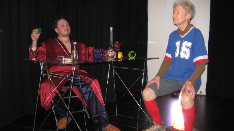 Zwei Personen in einem Raum. Eine Person tr&auml;gt einen roten Bademantel und h&auml;lt einen Zauberw&uuml;rfel. Die andere Person sitzt in Sportkleidung auf einem Verkehrskegel., &copy; Dreigroschentheater