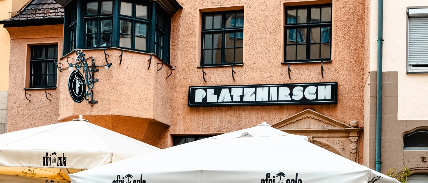 Gebäude mit dem Schild 'Platzhirsch', darunter Sonnenschirme mit 'afri cola'-Logo. Einladende Atmosphäre mit Speisekarten im Freien., © SMG Stuttgart Marketing GmbH - Sarah Schmid