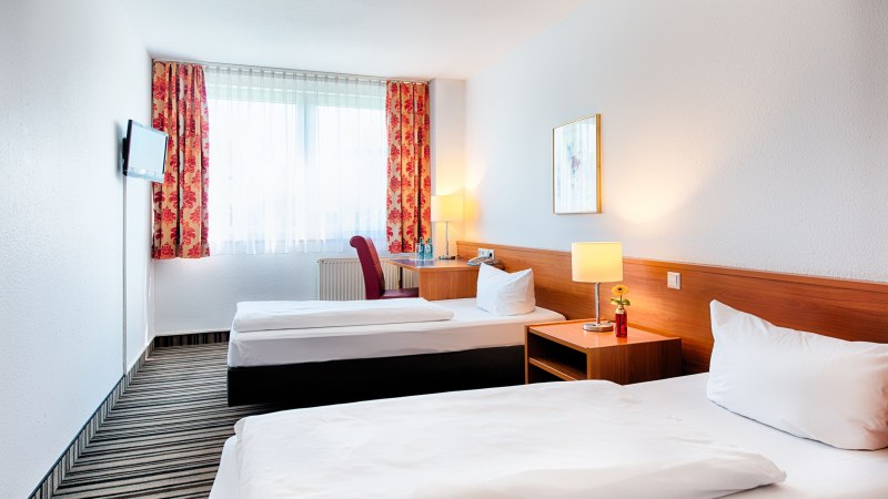 A bright twin room with two single beds, a nightstand with a lamp, a desk, a TV, and red curtains., &copy; ACHAT Hotels Deutschland