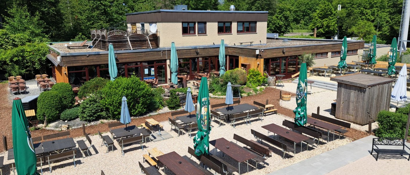 Außenbereich von Weitmanns Waldhaus mit Holztischen, Bänken und Sonnenschirmen, umgeben von grünen Bäumen und blauen Himmel., © Timo Weitmann Außenbereich von Weitmanns Waldhaus mit Holztischen, Bänken und Sonnenschirmen, umgeben von grünen Bäumen und blauen Himmel., © Timo Weitmann