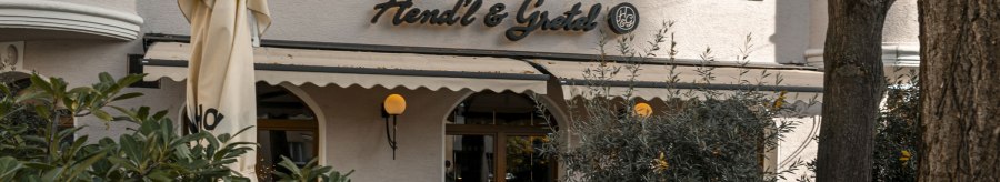Das Bild zeigt die Fassade des Restaurants Hendl & Gretel mit einem Sonnenschirm und Pflanzen im Vordergrund., © SMG, Sarah Schmid Das Bild zeigt die Fassade des Restaurants Hendl & Gretel mit einem Sonnenschirm und Pflanzen im Vordergrund., © SMG, Sarah Schmid