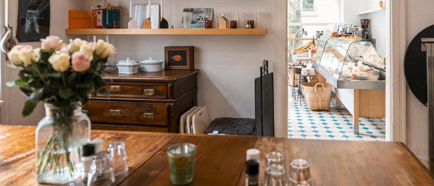 Ein gemütlicher Feinkostladen mit Holztisch, Blumenstrauß und einer Käsetheke im Hintergrund auf blau-weißem Fliesenboden., © SMG, Sarah Schmid