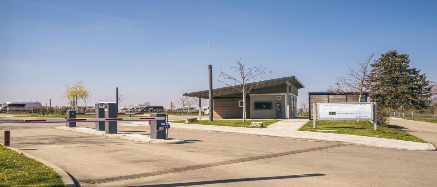 Einfahrt zum Wohnmobilstellplatz Beuren mit Schranke, kleinem Gebäude und Hinweisschild. Wohnmobile im Hintergrund, blauer Himmel., © SMG, Sarah Schmid