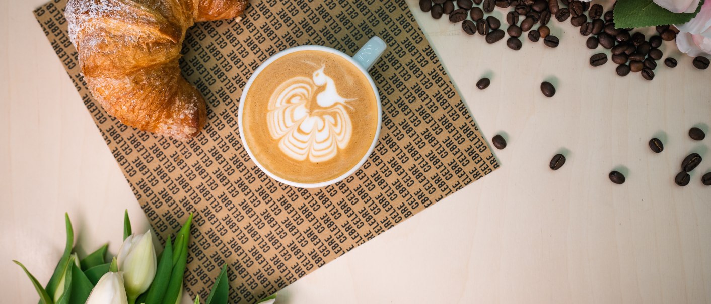 A croissant and cappuccino with latte art on a table surrounded by coffee beans, cotton, roses and tulips., © CELBRE