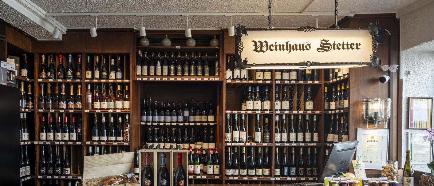 Counter at Weinhaus Stetter with shelves full of wine bottles and a wooden counter. A sign reading 'Weinhaus Stetter' hangs from the ceiling., © SMG Stuttgart Marketing GmbH - Sarah Schmid Counter at Weinhaus Stetter with shelves full of wine bottles and a wooden counter. A sign reading 'Weinhaus Stetter' hangs from the ceiling., © SMG Stuttgart Marketing GmbH - Sarah Schmid