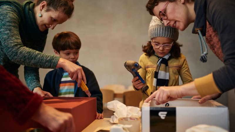 Zwei Erwachsene und zwei Kinder basteln an einem Tisch mit Kartons und Werkzeugen. Die Kinder halten Klebepistolen., &copy; Alex Wunsch