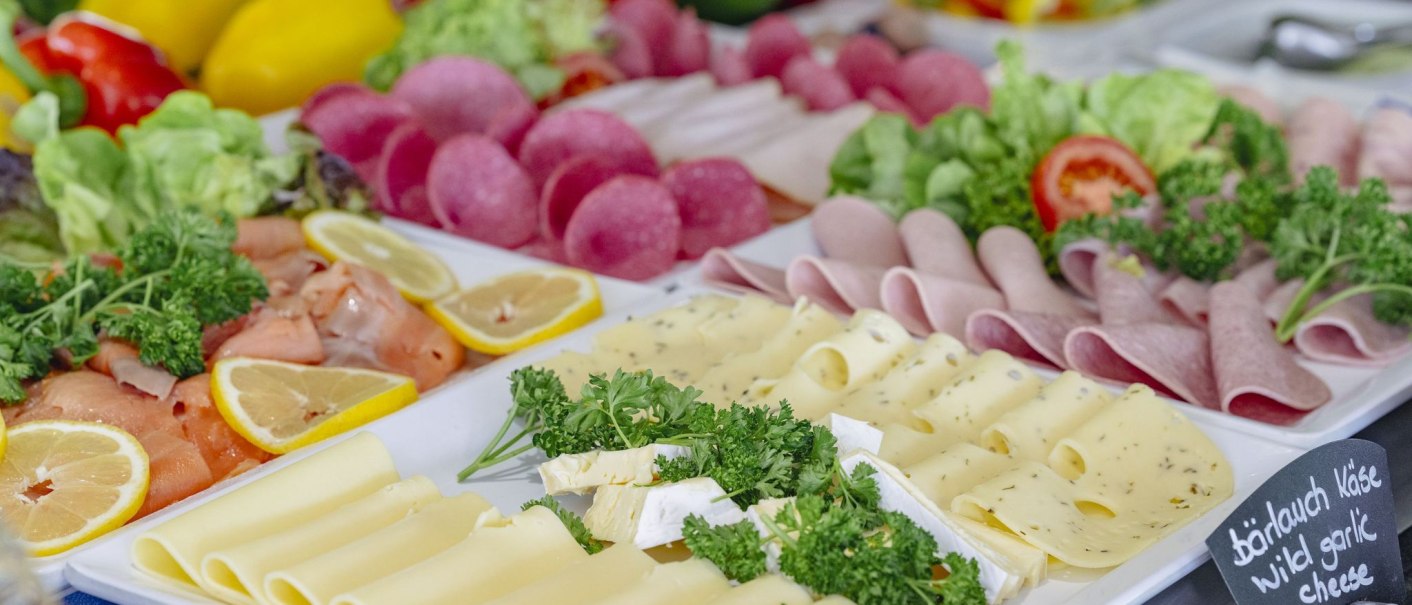 A breakfast buffet with various cheeses, sausage, salmon, lemon slices and fresh vegetables arranged on a table., © Plaza INN Stuttgart-Filderstadt A breakfast buffet with various cheeses, sausage, salmon, lemon slices and fresh vegetables arranged on a table., © Plaza INN Stuttgart-Filderstadt