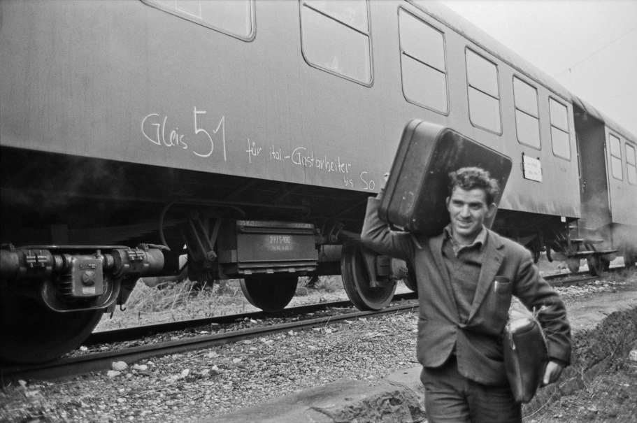 A man carries two suitcases next to a train with the inscription 'Gleis 51 für ital. Guest workers'., © Landesarchiv Baden-Württemberg HStAS Q 2/50 Nr. 2926-29 A man carries two suitcases next to a train with the inscription 'Gleis 51 für ital. Guest workers'., © Landesarchiv Baden-Württemberg HStAS Q 2/50 Nr. 2926-29