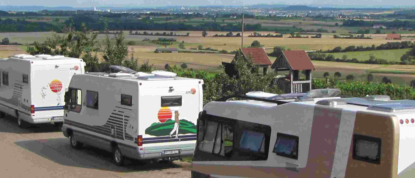 Wohnmobile parken auf einem Stellplatz mit Blick auf eine weite Landschaft und Felder. Im Hintergrund sind H&uuml;gel und eine Stadt zu sehen., &copy; PANORAMAWEINGUT Baumg&auml;rtner