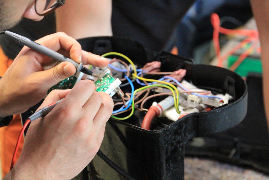 Nahaufnahme einer Person, die mit einem Lötkolben an einer Platine arbeitet, umgeben von bunten Kabeln., © Uprety 2024