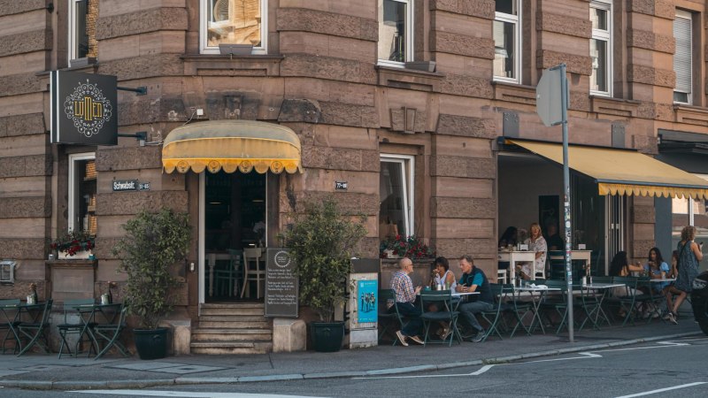 Café mit gelbem Vordach an einer Straßenecke. Gäste sitzen draußen an Tischen. Schild mit 'Lumen' und Straßenschild 'Schwabstr.' sichtbar., © SMG, Sarah Schmid
