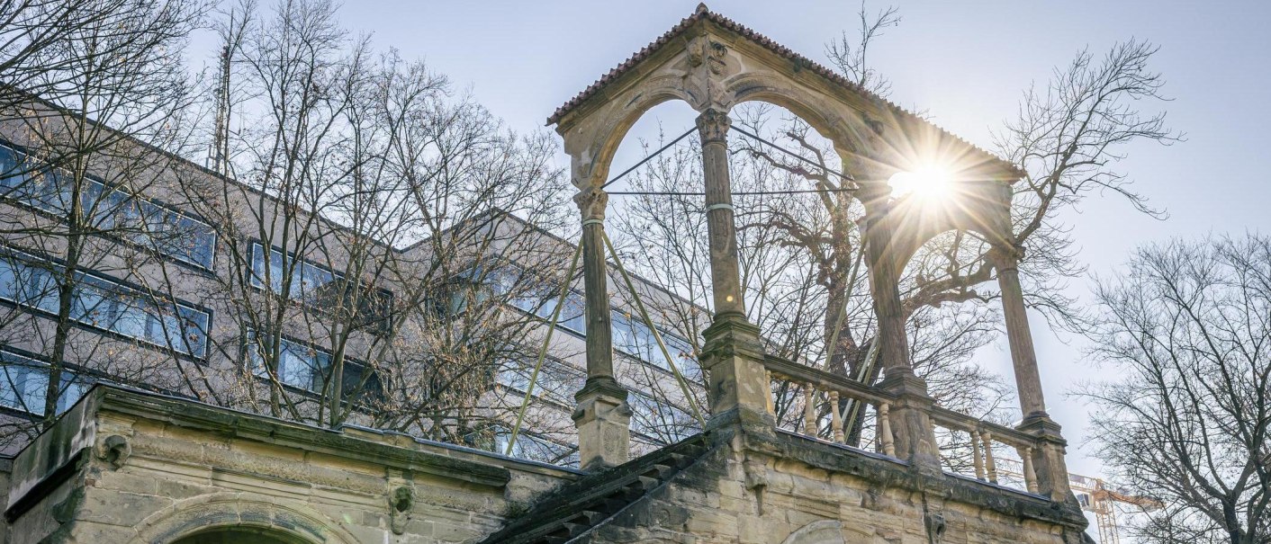 The ruins of the Neues Lusthaus with stone arches, surrounded by bare trees. Rays of sunlight break through the branches. A modern building in the background., &copy; Stuttgart-Marketing GmbH, Sarah Schmid