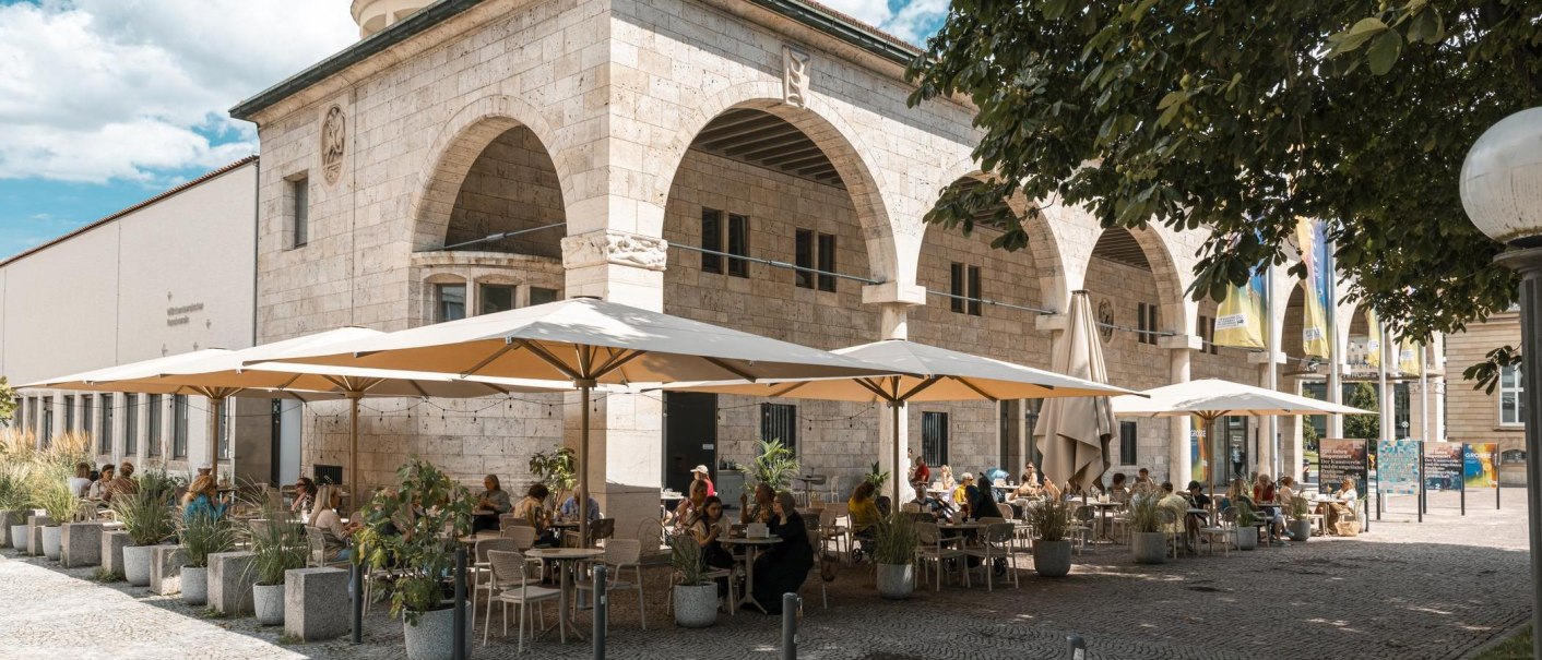 Ein Café mit großen Sonnenschirmen und Gästen, die draußen an Tischen sitzen. Das Gebäude hat Bögen und ist aus Stein., © Stuttgart Marketing GmbH, Sarah Schmid Ein Café mit großen Sonnenschirmen und Gästen, die draußen an Tischen sitzen. Das Gebäude hat Bögen und ist aus Stein., © Stuttgart Marketing GmbH, Sarah Schmid